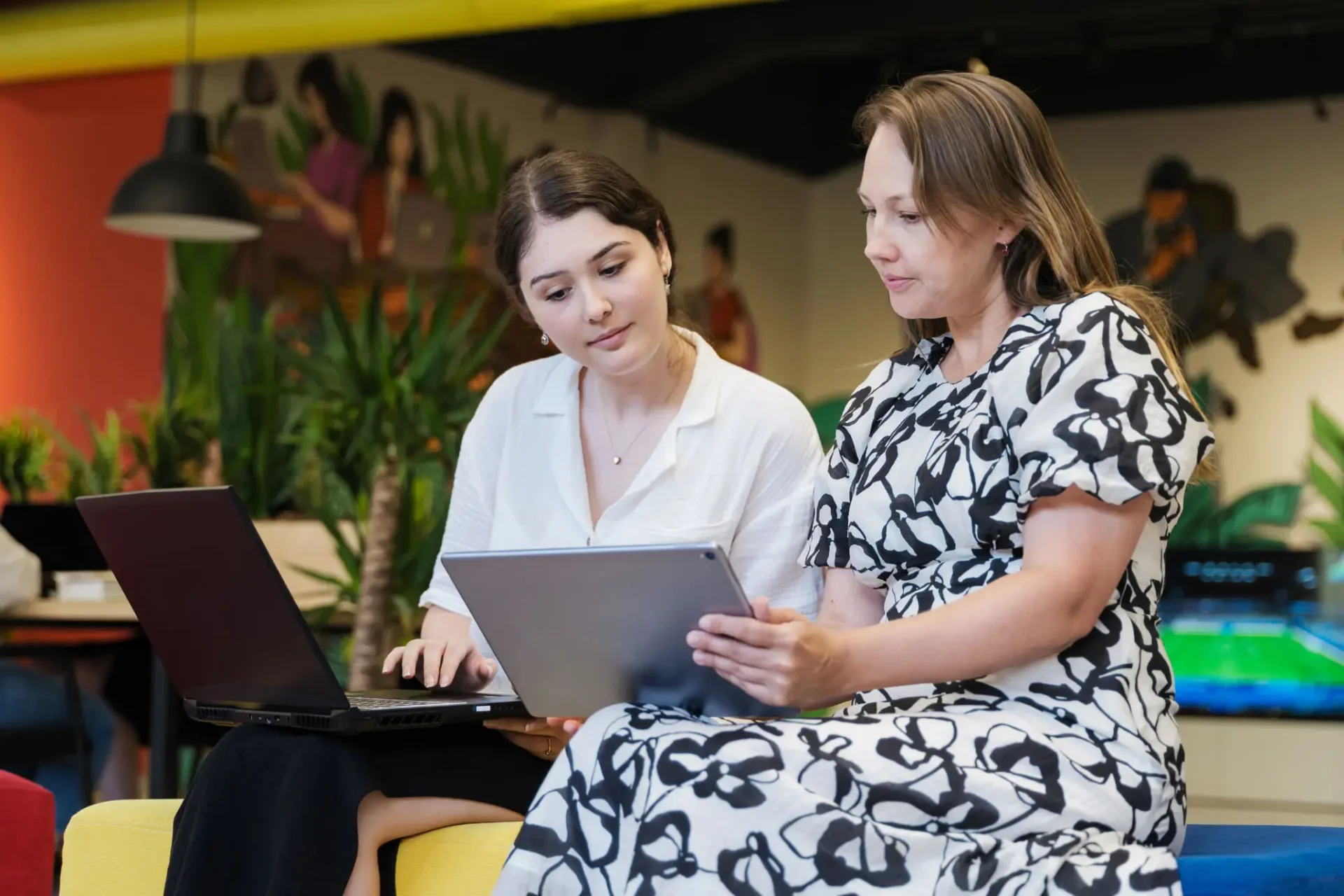 Two women collaborating with a laptop and tablet in a casual setting.