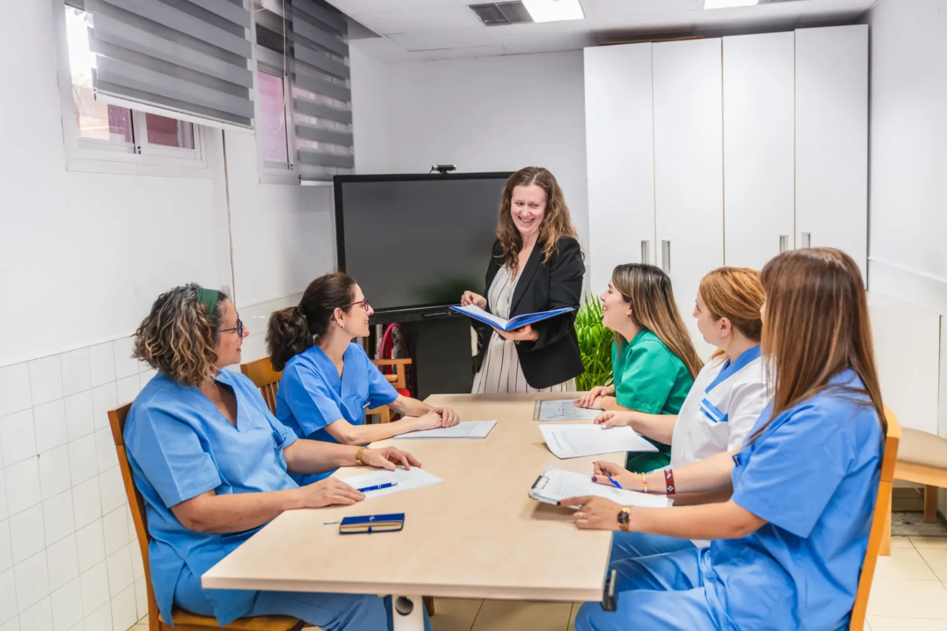 Healthcare professionals in scrubs attending a meeting with a standing presenter.